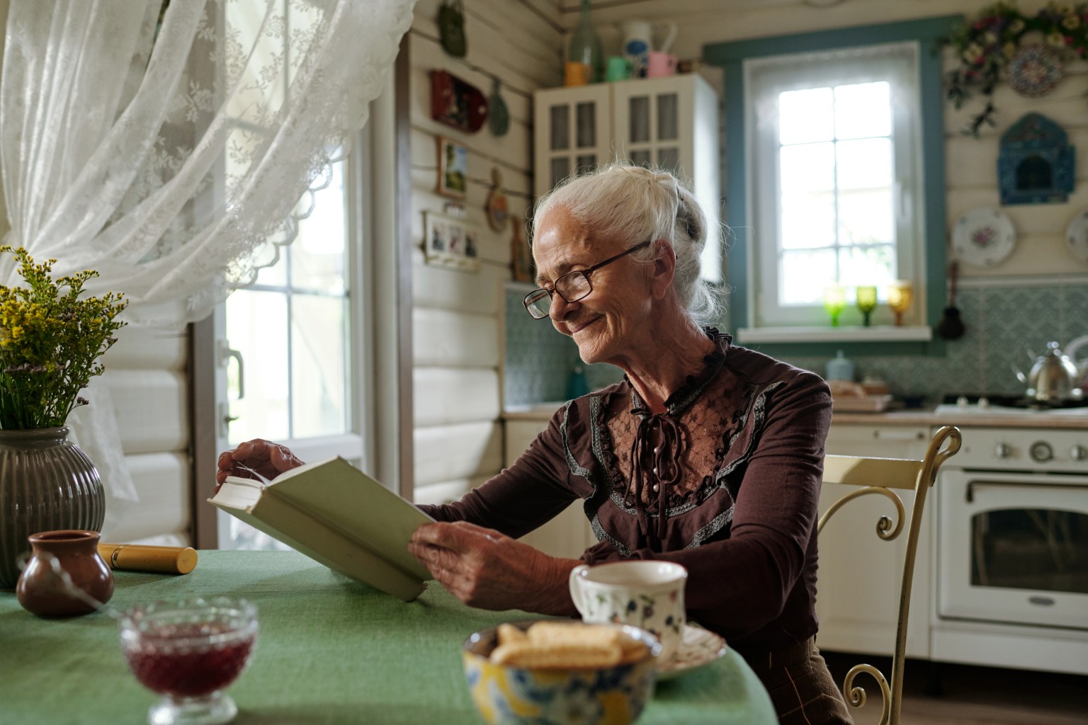 A woman sits reading a book at a kitchen table with a mug and a glass jar beside her in daylight.