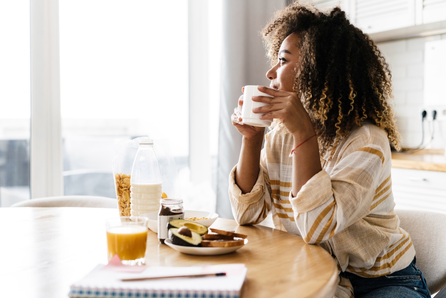 A woman sits at a round kitchen table by a bright window holding a mug, with juice, bread and a notebook in front of her.